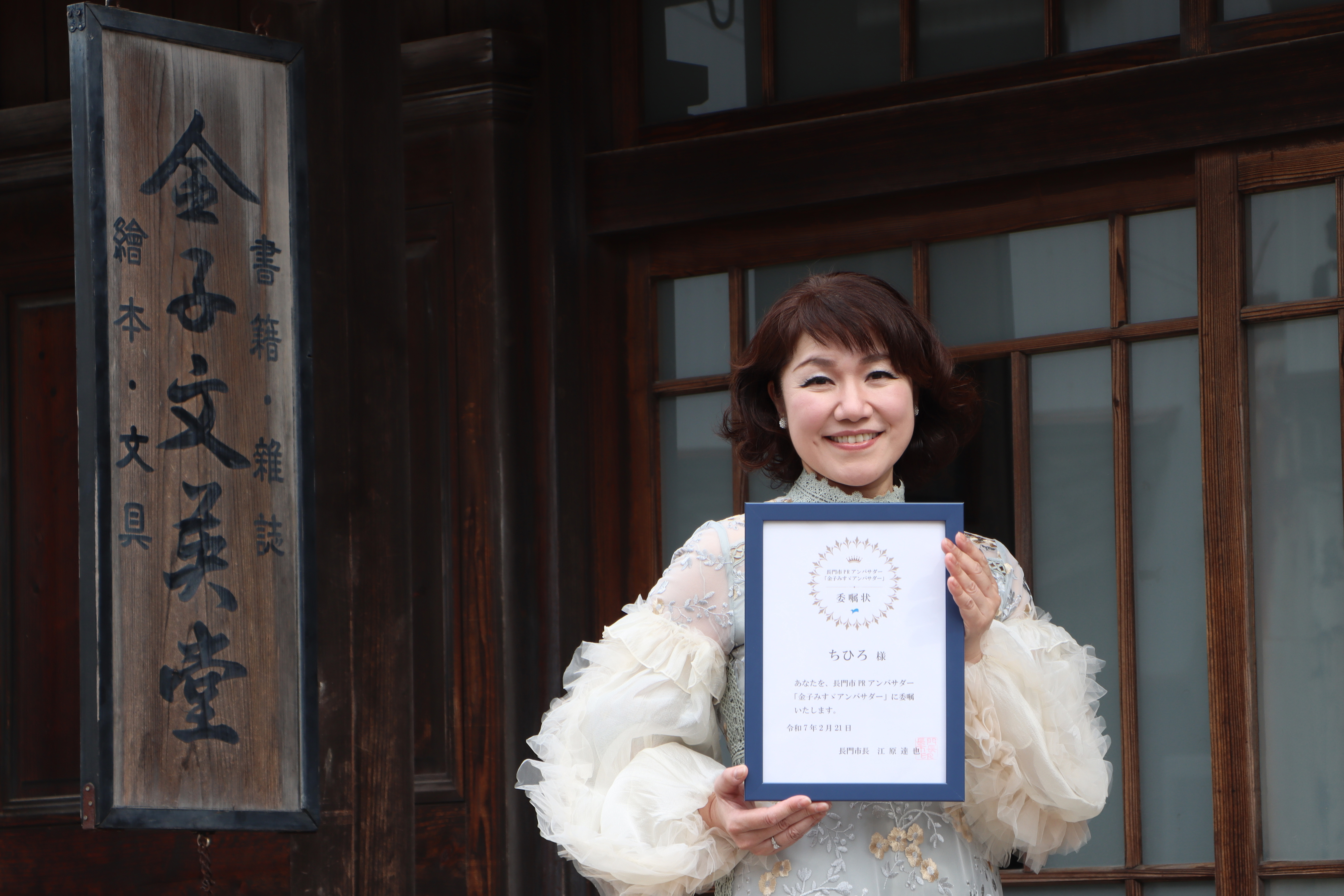 金子みすゞ記念館の前でみすゞアンバサダー就任の委嘱状を持つちひろさん